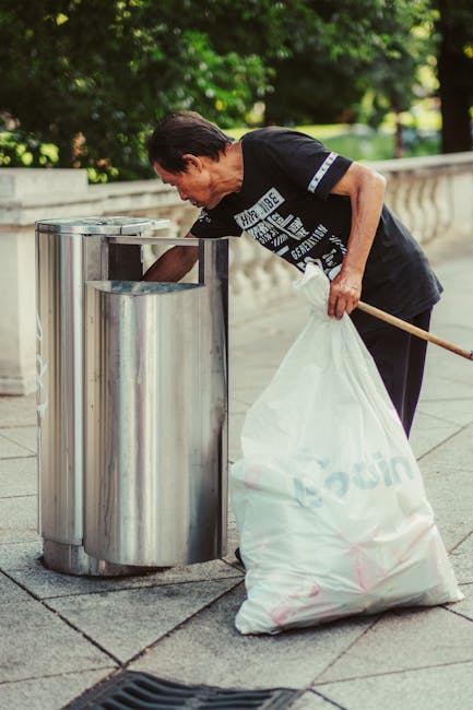 A man with short dark hair, wearing a black T-shirt with white text and dark trousers, is seen emptying a large white rubbish bag into a tall, cylindrical metal bin located on a paved sidewalk. The bin is made of brushed stainless steel, featuring a rounded top with an opening for waste disposal, positioned next to a low stone or concrete railing. In the background, there are lush green trees and a well-maintained park or green space, suggesting an outdoor urban environment. The man is leaning forward with his left hand gripping the bag and his right hand holding a wooden stick or broom handle, assisting him in managing the rubbish. The scene is lit with natural daylight, highlighting the reflective surface of the metal bin and the textures of the grey paving stones. This image from Rubbish Clearance Brompton illustrates on-site rubbish collection or waste removal service, fitting within the context of private disposal and alternative waste handling methods in a city park setting.