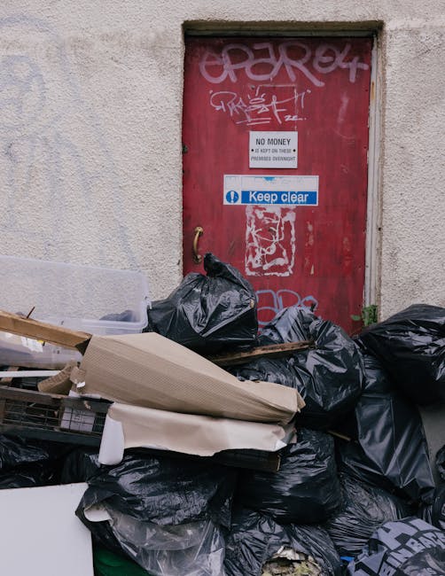 The image shows a large pile of black rubbish bags accumulated against a textured, off-white exterior wall beneath a small, rectangular door with a red, weathered surface. The door is covered with various graffiti tags in white and pink, with some overlapping. Several of the black plastic bags are stacked and leaning, with some appearing slightly torn or bulging, indicating recent or ongoing waste disposal. Among the rubbish are flattened cardboard boxes, one of which is light brown and ribbed in texture, partially unfolded and lying atop other bags; there are also some loose packaging and paper materials. Above the rubbish, attached to the door, is a white notice with black text stating 'NO MONEY' and a smaller 'Keep clear' sign in blue and white, the latter featuring a circular symbol. The scene suggests an area typical of alleyway or backstreet waste accumulation, which could relate to private waste handling services like those offered by Rubbish Clearance Brompton, providing an alternative to standard municipal rubbish collection.