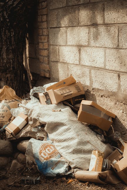 A pile of discarded rubbish situated at the base of a brick wall next to a tree, consisting of flattened cardboard boxes, crumpled paper bags, and a large woven sack. The cardboard boxes vary in size, with some partially opened or torn, revealing a grey interior, and display printed labels and barcodes. The paper bags are loosely crumpled, with a brown paper bag clearly visible among the debris. The woven sack appears worn, with some creases and dirt marks on its fabric surface. The surrounding environment suggests an outdoor area with uneven ground made up of soil and small rocks; the brick wall behind is weathered with visible mortar lines, and the scene is lit with natural daylight, providing a neutral, unobtrusive context that could relate to private waste disposal outside a property, as handled by services like Rubbish Clearance Brompton. This visual emphasizes the need for efficient rubbish removal and environmentally responsible waste handling methods, highlighting an inset of typical refuse that often requires professional clearance services for proper disposal.