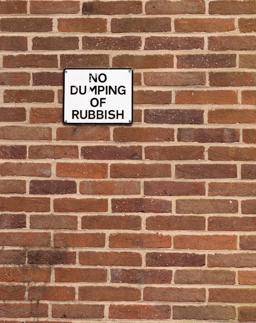 The image shows a large pile of black rubbish bags accumulated against a textured, off-white exterior wall beneath a small, rectangular door with a red, weathered surface. The door is covered with various graffiti tags in white and pink, with some overlapping. Several of the black plastic bags are stacked and leaning, with some appearing slightly torn or bulging, indicating recent or ongoing waste disposal. Among the rubbish are flattened cardboard boxes, one of which is light brown and ribbed in texture, partially unfolded and lying atop other bags; there are also some loose packaging and paper materials. Above the rubbish, attached to the door, is a white notice with black text stating 'NO MONEY' and a smaller 'Keep clear' sign in blue and white, the latter featuring a circular symbol. The scene suggests an area typical of alleyway or backstreet waste accumulation, which could relate to private waste handling services like those offered by Rubbish Clearance Brompton, providing an alternative to standard municipal rubbish collection.