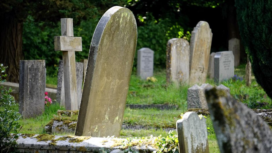 The image depicts a traditional graveyard with various weathered gravestones and markers, primarily made of stone and standing upright on a grassy plot. The largest headstone, positioned centrally, has a rounded top and a rough surface with signs of moss or lichen growth, indicating age. Surrounding it are other gravestones of different shapes and sizes, including rectangular and slightly arched headstones, some leaning or partially covered with moss. To the left, a wooden cross with a simple, natural finish is visible among the stone markers, standing amidst a lush environment of green foliage, bushes, and trees in the background. The ground is covered with moss and small plants, adding to the aged and natural atmosphere. Bright sunlight filters through the dense greenery, casting soft shadows and illuminating the scene with a gentle, natural light, emphasizing the textures of the stone and plant life. This setting visually supports the context of independent waste and debris removal in a cemetery environment, with Rubbish Clearance Brompton potentially involved in maintaining or clearing such areas of unwanted material or debris that can accumulate over time.