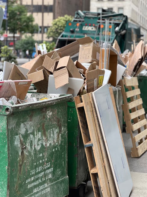 A large green commercial waste skip filled with a mixture of cardboard boxes, some flattened and others slightly crumpled, along with a few white packaging materials and smaller debris. The boxes vary in size and are made from plain brown or white cardboard, with some showing shipping labels or tape. Leaning against the side of the skip is a wooden pallet with a metal strip, possibly part of furniture or shelving, partially visible and extending towards the right of the image. In the background, a green waste collection truck equipped for rubbish removal is parked on a city street lined with office or apartment buildings, some trees, and parked cars. The sky appears overcast, providing diffuse lighting over the scene. The scene suggests an instance of private waste disposal or on-site clearance, typical of independent rubbish collection services such as those offered by Rubbish Clearance Brompton, rather than state-provided rubbish collection, with the focus on visible waste materials ready for removal or disposal.
