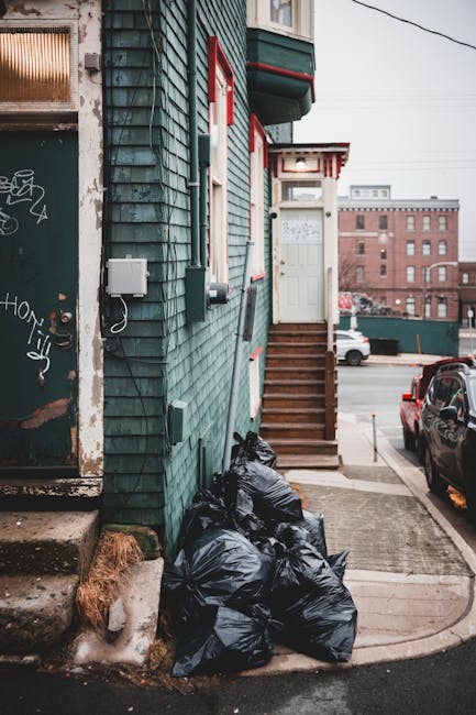 A close-up view of a residential building with dark green wooden cladding showing weathered and chipped paint, adjacent to a sidewalk. Several black rubbish bags are piled at the bottom of a set of outdoor wooden stairs leading up to a white door with red trim and a small porch roof. The bags appear to contain waste materials and are placed directly on the pavement, with some leaning against the building's wall. To the left, part of another door or window with graffiti and rusted metal elements is visible. In the background, an urban street scene with parked cars, a red brick building, and overhanging power lines can be seen, indicating a typical city environment. The scene is lit with natural daylight, emphasizing the textures of the building's siding and the trash bags, illustrating a typical example of private rubbish collection or alternative waste handling undertaken by a local waste removal service like Rubbish Clearance Brompton.