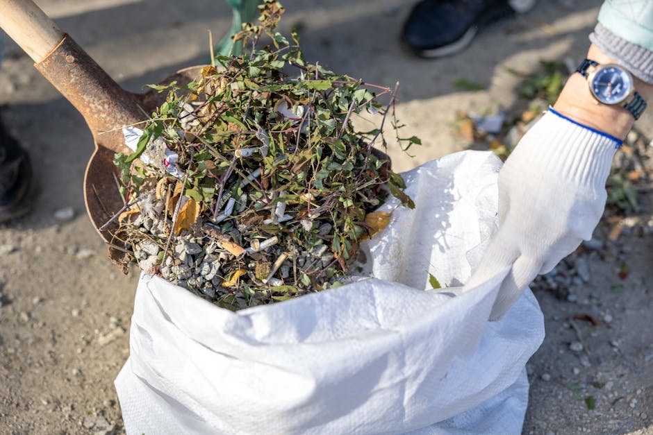 A gloved hand is holding open a white woven polypropylene sack filled with a mixture of greenish-brown dried plant material, small broken branches, and scattered gravel or rubble, placed on a paved outdoor surface. A rusty shovel with a wooden handle is positioned nearby, partly obscured, as if in the process of loading or lifting waste. In the background, a pair of black shoes and a person's wrist wearing a blue watch are visible, suggesting a person's presence during a rubbish clearance operation. The scene appears to be taking place outside, possibly on a driveway or garden path, with natural daylight illuminating the clutter and providing clear detail of the collected debris. This image exemplifies a collection of garden or yard waste ready for disposal, typical of private waste handling or independent rubbish removal services, like those offered by Rubbish Clearance Brompton, highlighting the process of clearing organic and mixed waste.