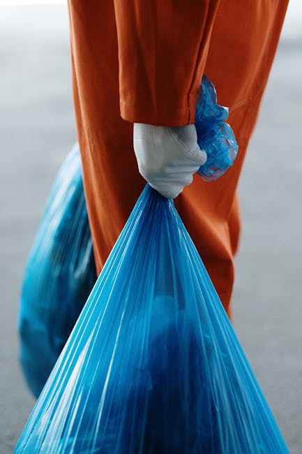 A gloved hand is holding open a white woven polypropylene sack filled with a mixture of greenish-brown dried plant material, small broken branches, and scattered gravel or rubble, placed on a paved outdoor surface. A rusty shovel with a wooden handle is positioned nearby, partly obscured, as if in the process of loading or lifting waste. In the background, a pair of black shoes and a person's wrist wearing a blue watch are visible, suggesting a person's presence during a rubbish clearance operation. The scene appears to be taking place outside, possibly on a driveway or garden path, with natural daylight illuminating the clutter and providing clear detail of the collected debris. This image exemplifies a collection of garden or yard waste ready for disposal, typical of private waste handling or independent rubbish removal services, like those offered by Rubbish Clearance Brompton, highlighting the process of clearing organic and mixed waste.